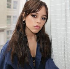 a woman with long brown hair wearing a dark blue shirt and earrings sitting in front of a window