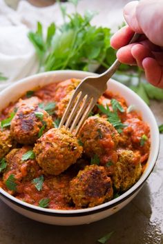 a hand holding a fork over a bowl of meatballs with sauce and parsley