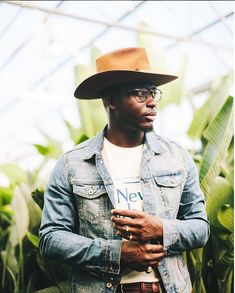 a man wearing a hat and glasses standing in front of some plants with his hands on his hips