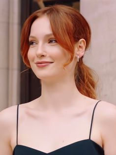 a woman with red hair wearing a black dress and smiling at the camera while standing in front of a building