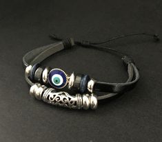 "This cute, casual, evil eye bracelet is a great gift item, stocking stuffer or a \"wear it everyday\" item for yourself! Two separate leather bands are tied together with a slide knot making it easily adjustable. The beads are a mix of metal, wood and plastic with an enamel eye. It's unusually light, making it very comfortable to wear." Trendy Adjustable Evil Eye Bracelet, Adjustable Spiritual Leather Bracelet For Friendship, Adjustable Evil Eye Bracelet For Friendship, Casual Adjustable Evil Eye Bracelet As Gift, Adjustable Silver Bohemian Evil Eye Bracelet, Bohemian Style Evil Eye Bracelet With Adjustable Cord, Adjustable Nickel-free Black Leather Bracelet, Adjustable Black Nickel-free Leather Bracelet, Trendy Adjustable Leather Bracelet For Festivals