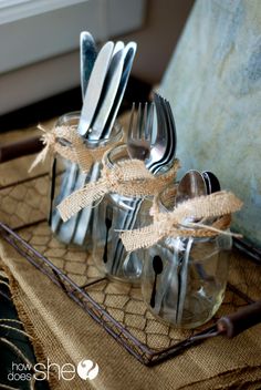 there are forks, spoons and knives in glass containers on a tray with burlap