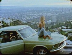 a woman sitting on the hood of a green car in front of a cityscape