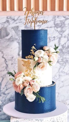 a three tiered blue and white cake with flowers on the top is displayed in front of a marble wall