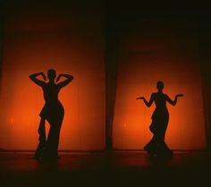 three silhouettes of two women dancing in front of an orange wall with lights behind them