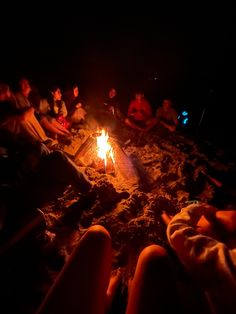 people sitting around a campfire at night on the beach with their feet in the sand