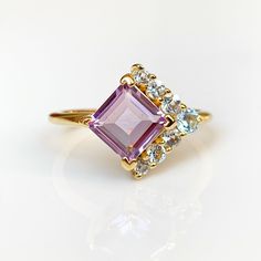 Indulge in affordable luxury with our square amethyst ring, a modern take on a timeless design. This captivating gold vermeil ring seamlessly combines ethically sourced, recycled silver with a alternative arrangement of amethyst and pale blue topaz stones. This piece was crafted with minimal environmental impact. As the birthstone for February, amethyst is celebrated for its calming properties, offering clarity in the midst of chaos. Embrace a sense of tranquility with this stunning ring that beautifully captures the essence of calm. Versatile enough for any occasion, this handcrafted 14ct gold vermeil ring comes in an elegant black gift box adorned with our gold foil logo--a perfect gift for a loved one or a treat for yourself. Handcrafted in 14ct gold vermeil Square-cut 7mm amethyst gems Luxury Amethyst Halo Ring For Gift, Square Halo Ring, Gold Foil Logo, Blue Topaz Stone, Amethyst Gem, Purple Band, Black Gift Boxes, Topaz Stone, Recycled Silver