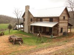 an old log cabin sits in the middle of a grassy field next to a wooden wagon