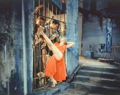 a woman in an orange dress leaning on a jail cell door with two men behind her