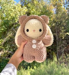 a hand holding a small stuffed animal in front of some trees and grass with one eye open