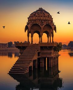 a gazebo sitting on top of a body of water at sunset with birds flying over it