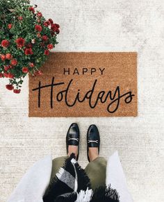 a person wearing black shoes standing in front of a happy holidays door mat with red flowers