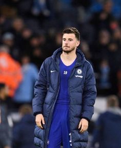 a man standing on top of a soccer field wearing a blue jacket and pants with his hands in his pockets