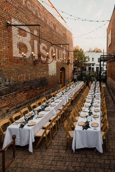 an outdoor dining area with long tables and white tablecloths set up in front of a brick building