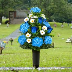 blue and white flowers in a black vase on the grass near gravestones at an outdoor cemetery