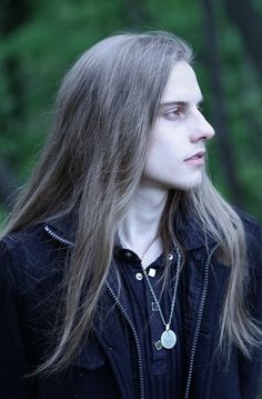 a woman with long hair wearing a black shirt and silver necklace standing in front of trees