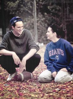 a man and woman sitting on the ground with leaves all around them, smiling at each other