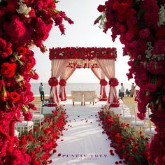 an outdoor ceremony setup with red flowers and white drapes on the aisle to the beach