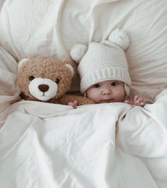 a baby laying in bed next to a teddy bear wearing a knitted white hat