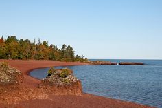 a body of water with trees in the background and red sand on the beach near to it