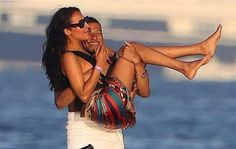 a man is holding a woman on his back while she rides a motorcycle by the water