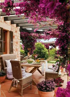 an outdoor dining area with wicker chairs and purple flowers on the arbored patio