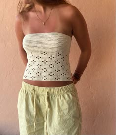 a woman standing in front of a pink wall wearing yellow shorts and a white crochet top