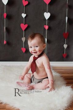 a baby wearing a tie and sitting on a rug with hearts hanging from the ceiling