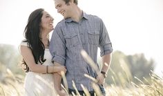 a young man and woman standing in tall grass smiling at each other while looking into the distance