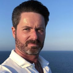 a close up of a person with a beard and white shirt near the ocean on a sunny day