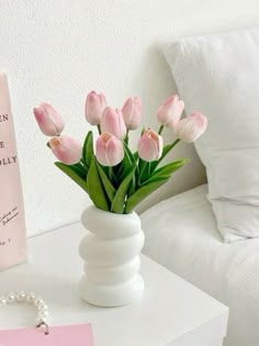 a white vase filled with pink tulips on top of a table next to a book