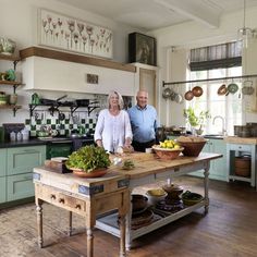 an older couple standing in the kitchen preparing food on a large wooden table with bowls and pans