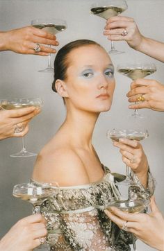 a woman with blue makeup holding wine glasses in front of her face and several other hands around her head