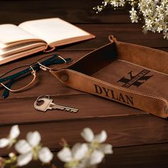 a leather case with glasses and keys on top of a wooden table next to an open book