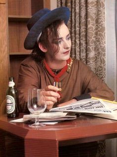 a man in a hat sitting at a table with a wine glass and newspaper on it