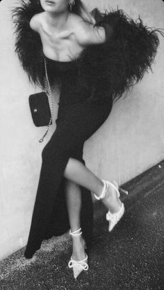 a woman leaning against a wall wearing a long dress and feathered jacket with her legs crossed