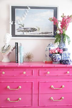 a pink dresser with flowers on top and pictures hanging above it in a room that has white walls