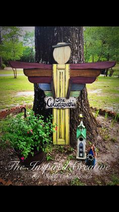 a wooden sign sitting next to a tree in the grass with flowers and plants around it