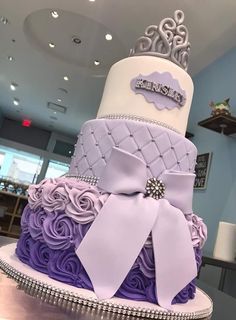 a three tiered cake decorated with purple and white ribbons, bows and jewels on top