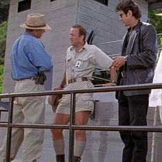 three men in shorts and hats standing on a balcony talking to each other while another man stands nearby