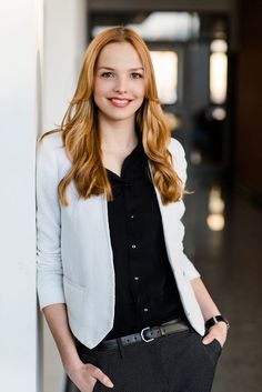 a woman standing next to a wall with her hands in her pockets and looking at the camera