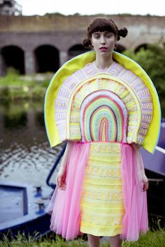 a woman standing in front of a body of water wearing a colorful dress and large fan