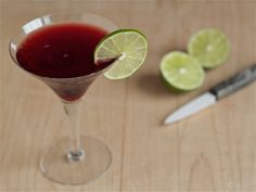 a martini glass filled with red liquid and limes next to a knife on a wooden table