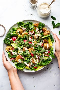 two hands holding a plate of salad with dressing on the table next to some ingredients