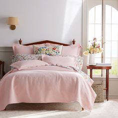 a bed with pink linens and pillows in a white room next to a window