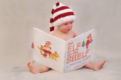 a baby sitting on the floor while reading a book with a red and white knitted hat