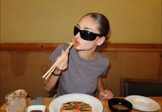 a woman sitting at a table with chopsticks in her mouth and eating pizza