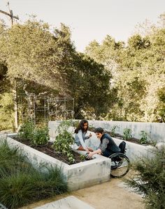 two people sitting in an outdoor garden with plants and flowers on the ground next to each other