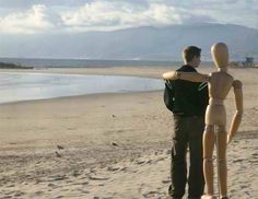 a man standing next to a wooden mannequin on a beach near the ocean