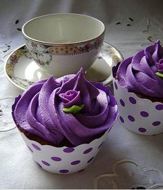 two cupcakes with purple frosting sitting on a table next to a teacup